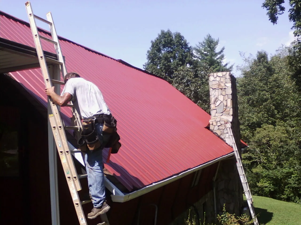 Roofer on a ladder beside a red metal roof and stone chimney — Soffit & Fascia Repair by Roofing Brothers in Campbellsport